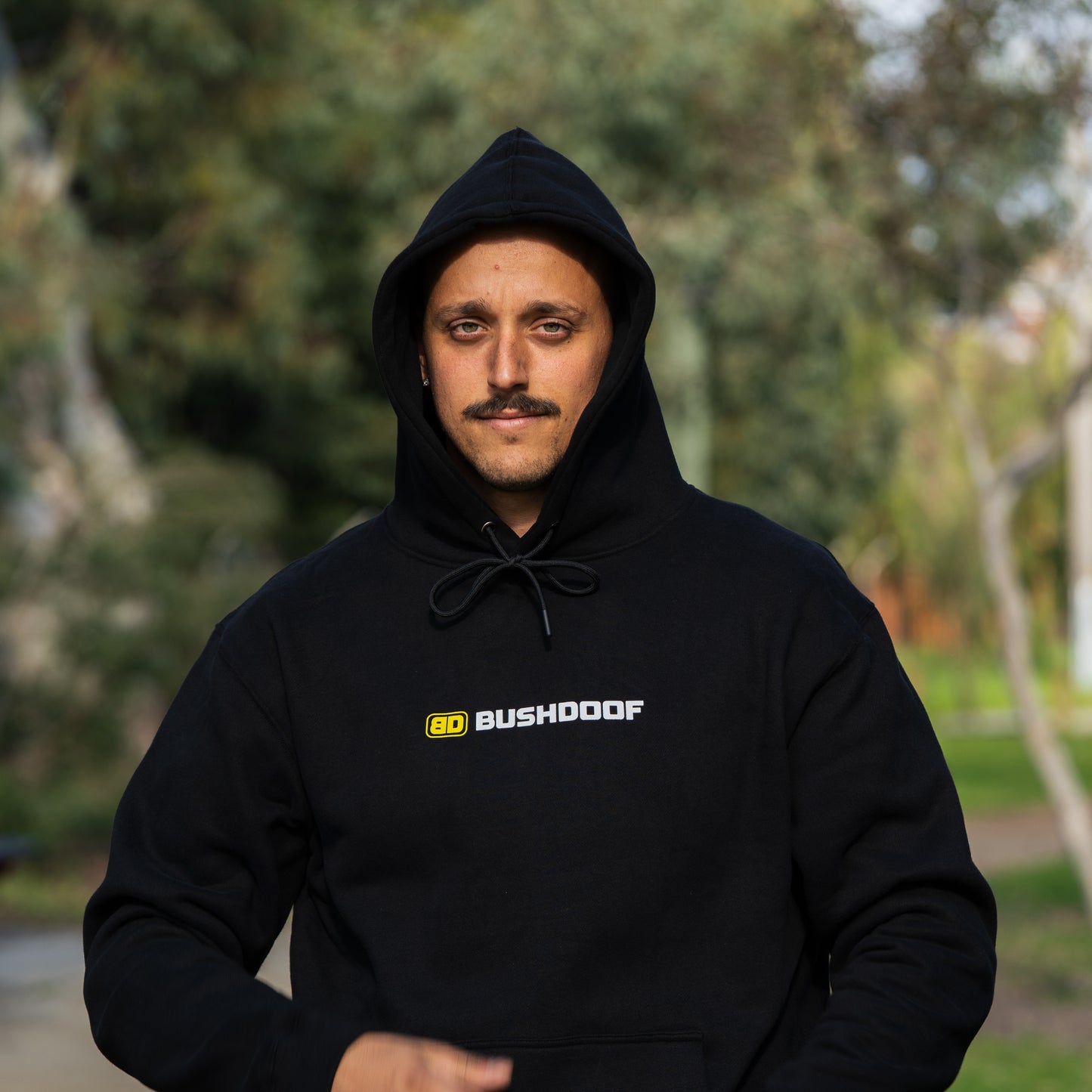 Bushdoof Staple Hoodie in black, hood up, worn outdoors